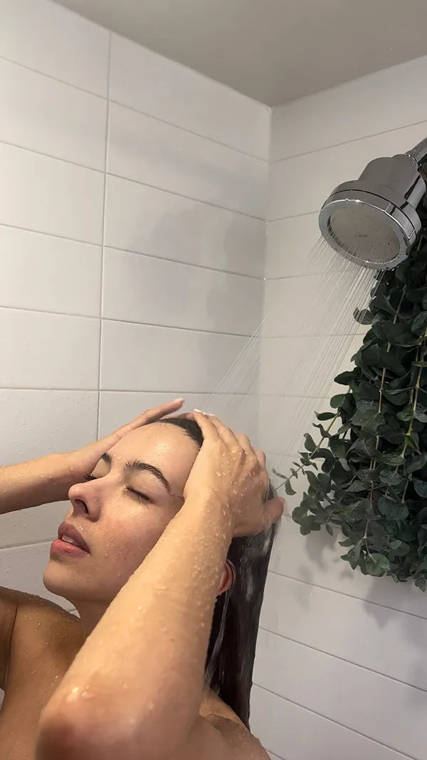 Person washing their hair under a shower with white tiles and green plant in the background.
